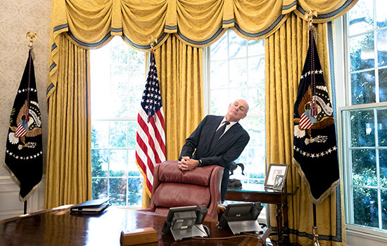Man leaning and looking off while standing behind a chair in the Oval Office.