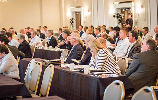 people at a conference sitting at tables. Camera people at the back of the room.